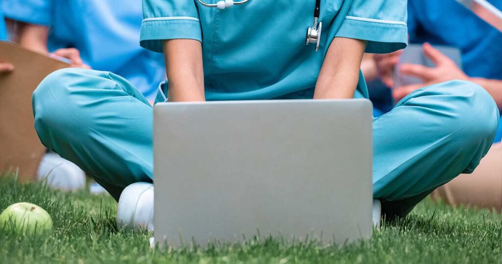 A nurse is continuing their education online using a laptop outside on the grass
